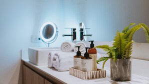Un tocador de baño con espejo, lavabo blanco y una planta en un florero. Hay toallas enrolladas y dos botellas de loción en la encimera.