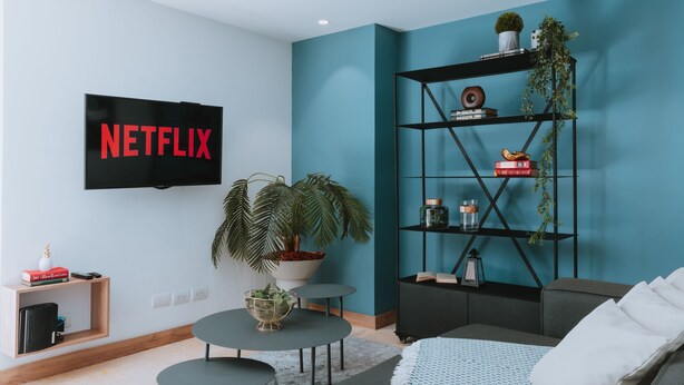 A modern living room with a teal accent wall, a mounted TV displaying "NETFLIX", a black bookshelf, a grey sofa, and a round coffee table.