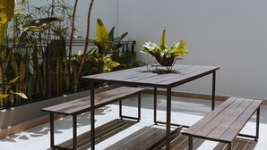 A patio with a wooden table and benches, surrounded by tropical plants and a metal fence.
