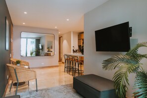 A modern living room with a large mirror, a flat-screen TV mounted on the wall, a seating area with a bench and chairs, and a kitchen area with bar stools. - Comfortable 895-foot apartment - 24/7 front desk - Alori 201 (Medellín)