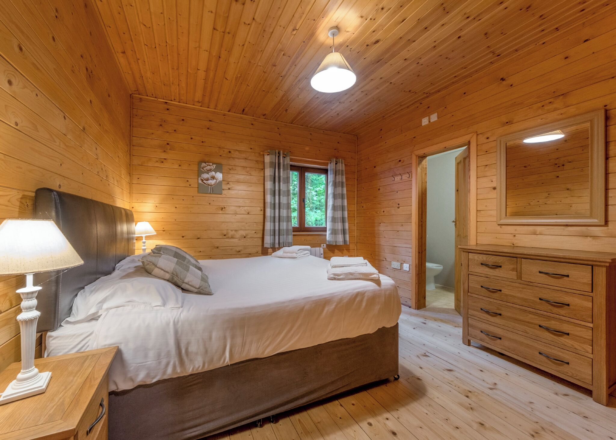 A wooden cabin bedroom with a bed, bedside lamps, a dresser, and a window with curtains.