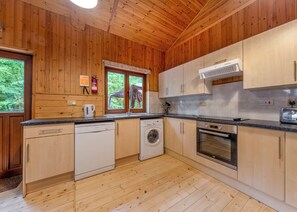 Cucina di una baita in legno con lavastoviglie, lavatrice, forno e una finestra vista verde.