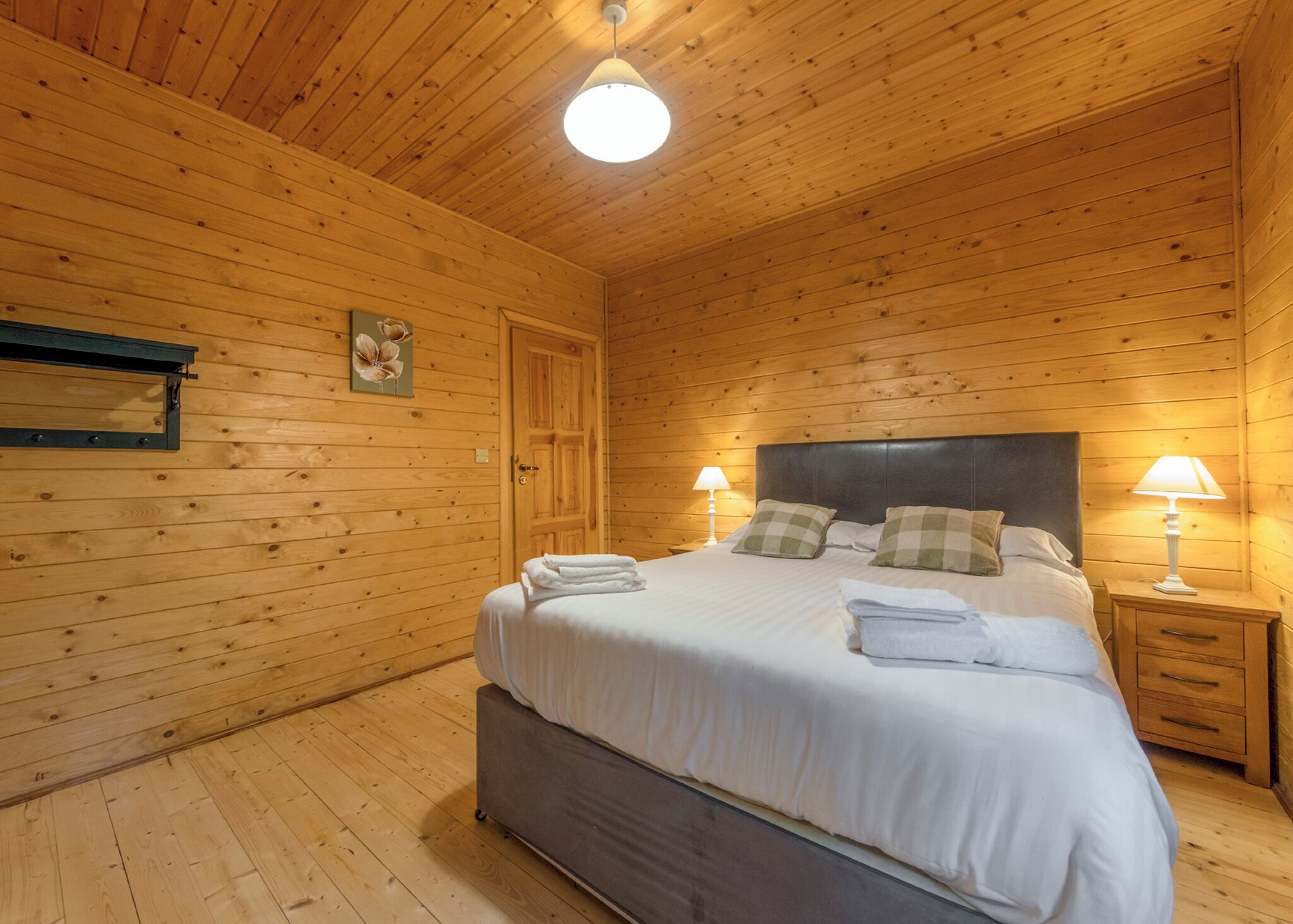 A wooden-paneled bedroom with a bed, two bedside tables with lamps, a door, and a wall-mounted shelf.