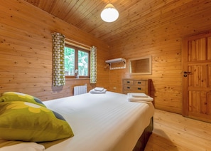 A wooden cabin room with a bed, a window with curtains, a radiator, a mirror, and a door.