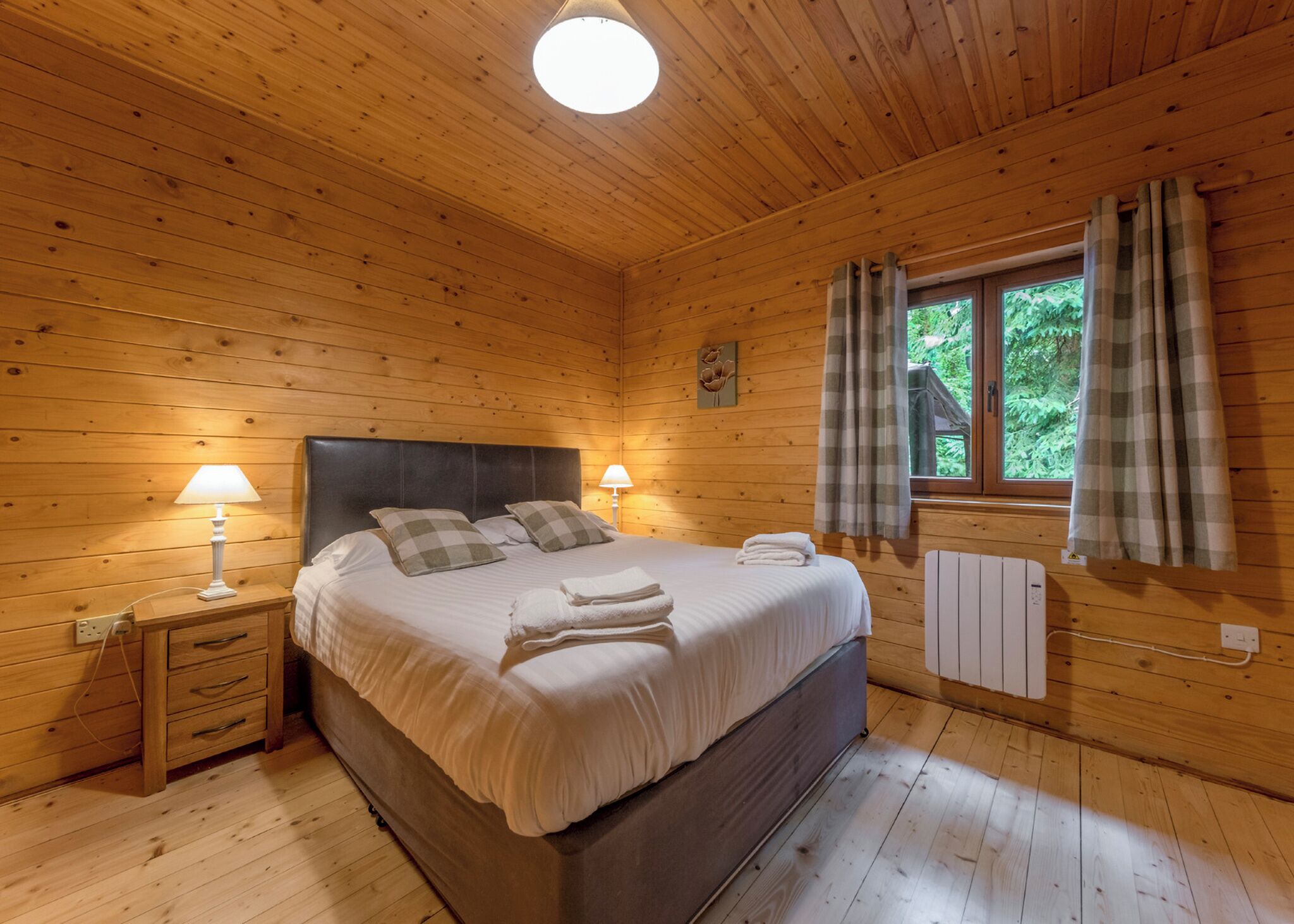 A wooden cabin bedroom with a bed, bedside lamps, a window with curtains, and a radiator.