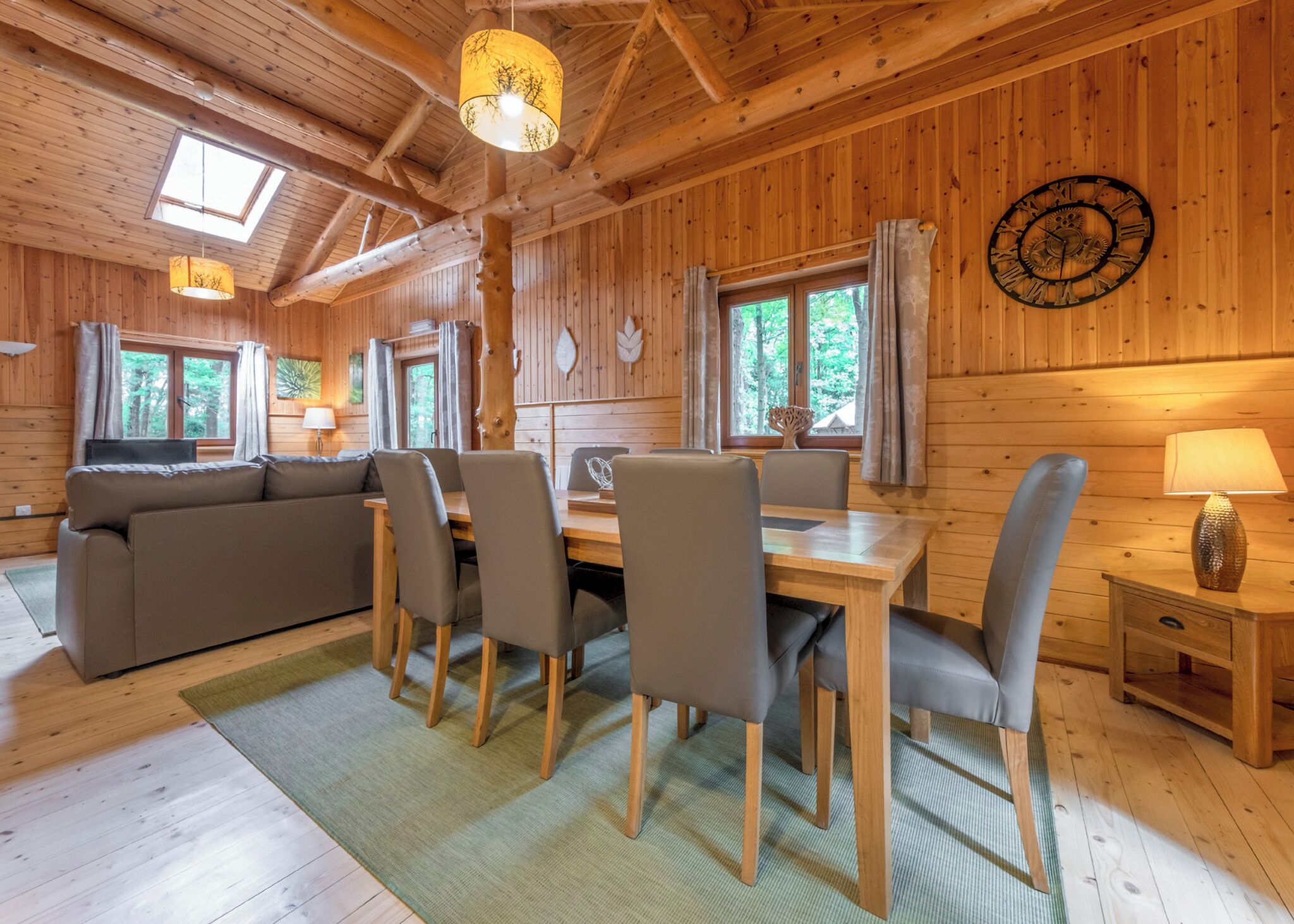 A wooden cabin interior with a dining table, chairs, a sofa, a clock, and a window with curtains.