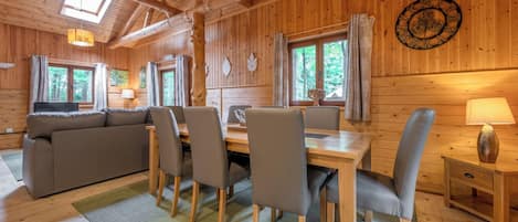 A wooden cabin interior with a dining table, chairs, a sofa, a clock, and a window with curtains.