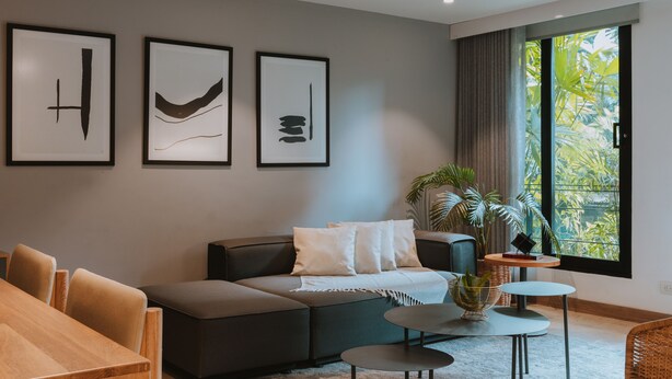 Ein modernes Wohnzimmer mit einer Couch, einem Couchtisch und einem Esstisch. Drei gerahmte Kunstwerke sind an der Wand zu sehen und ein großes Fenster bietet Blick ins Grüne.