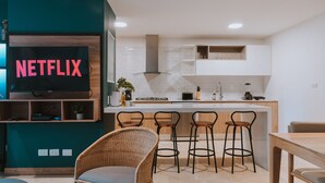 Eine moderne Küche mit integrierter Kochinsel, Barhocker, einem Flachbildfernseher, der 'NETFLIX' anzeigt, und einem Essbereich mit einem Korbstuhl.