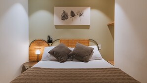 Uma cama bem arrumada com duas almofadas, uma obra de arte na parede com folhas, um abajur e uma pequena mesa com luminária.