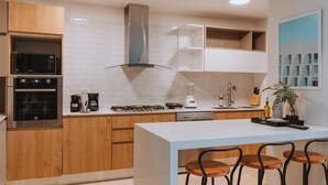 Cozinha moderna com armários de madeira, micro-ondas embutido, forno e lava-louças. Há uma ilha branca com banquetas e uma janela grande sobre a pia.