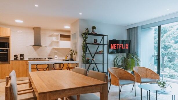 A modern kitchen with wooden cabinets, a built-in oven, and a dining area with a wooden table and wicker chairs. A large TV screen displays the Netflix logo. - 2 bedroom apartment in medellãn (Medellín)