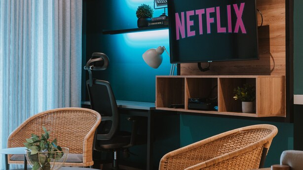 Una sala moderna con un televisor de pantalla plana grande mostrando "NETFLIX", sillas de mimbre, una mesita y un escritorio con una computadora.
