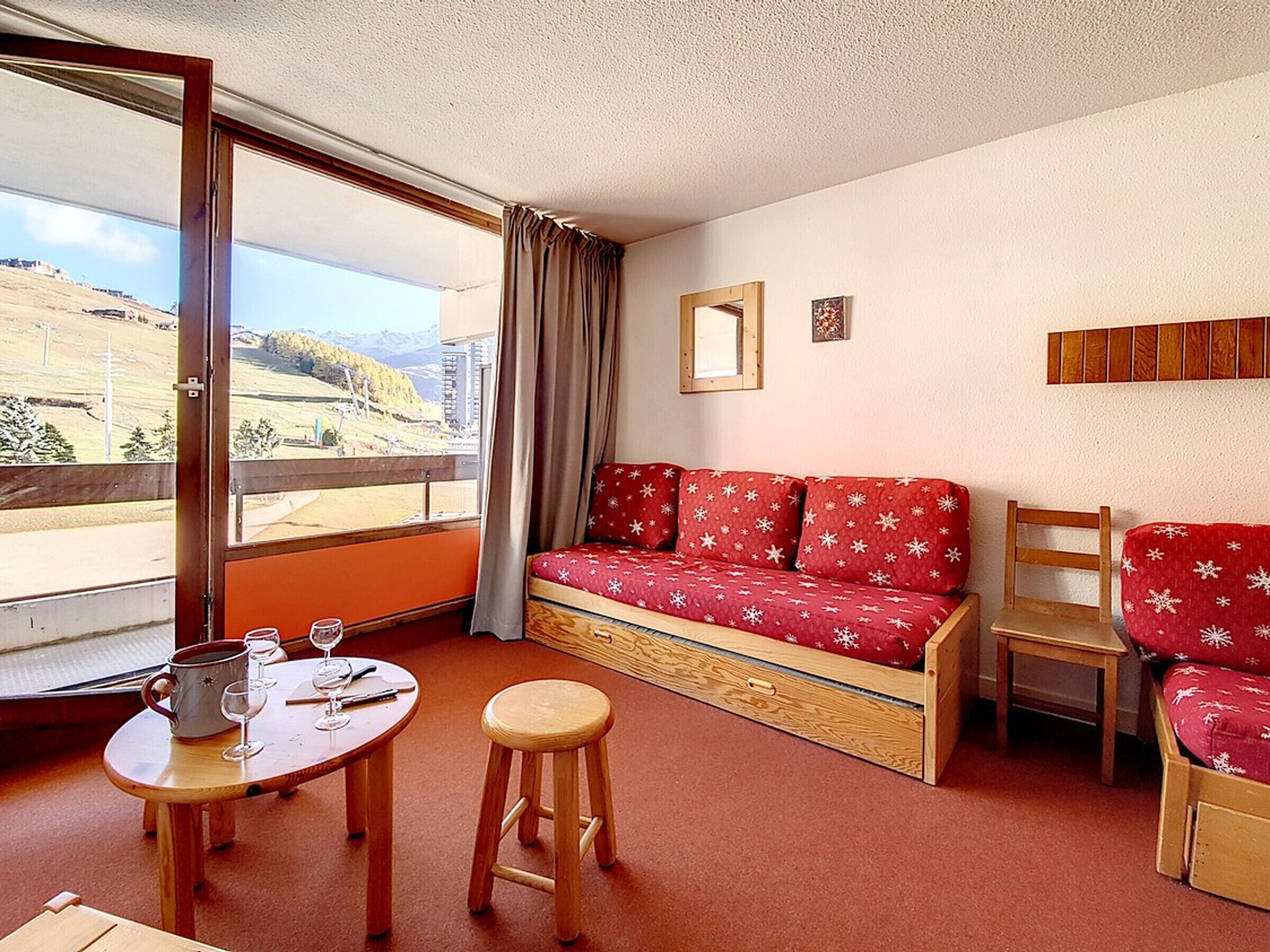 A room with a red sofa, a wooden chair, a small table with a stool, a window with a view of a landscape, and a mirror on the wall.