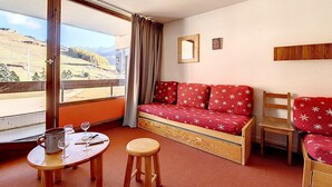 Ein Zimmer mit einem roten Sofa, einem Holzsessel, einem kleinen Tisch mit einem Hocker, einem Fenster mit Landschaftsblick und einem Spiegel an der Wand.