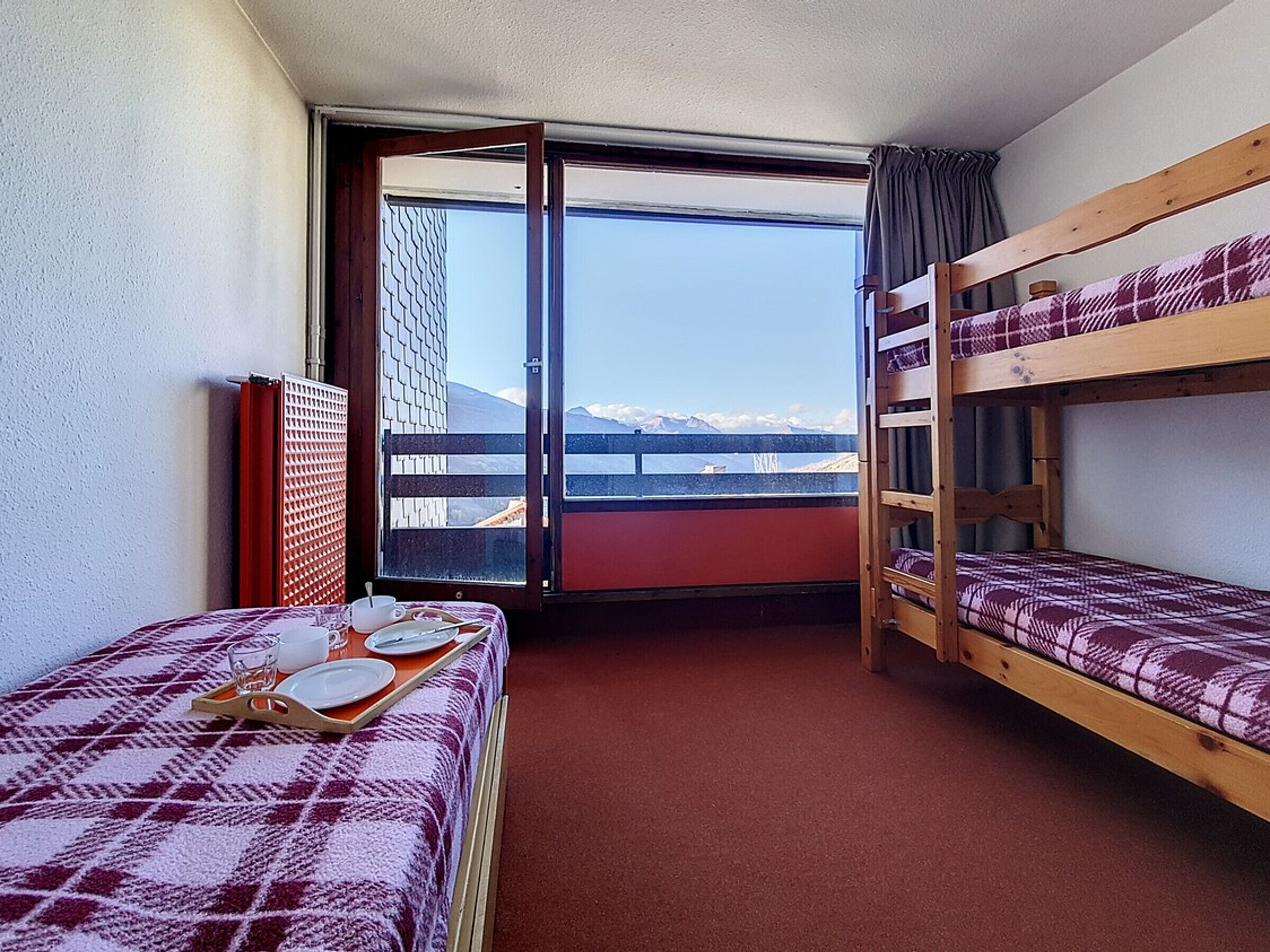 Una habitación pequeña y funcional con dos literas y una cama individual, una ventana con vistas a montañas y una alfombra roja.