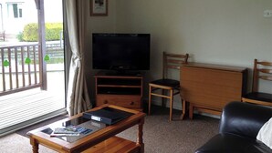 A living room with a television, wooden furniture, a small table, and a view through a glass door. - 2 bedroom accommodation in Wroxham, Norfolk Broads (Wroxham, Norfolk Broads)