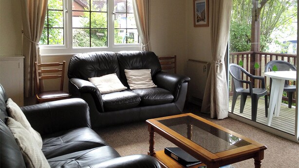 A living room with a black leather sofa, a glass coffee table, and a view of a garden through a sliding door. - 2 bedroom accommodation in Wroxham, Norfolk Broads (Wroxham, Norfolk Broads)