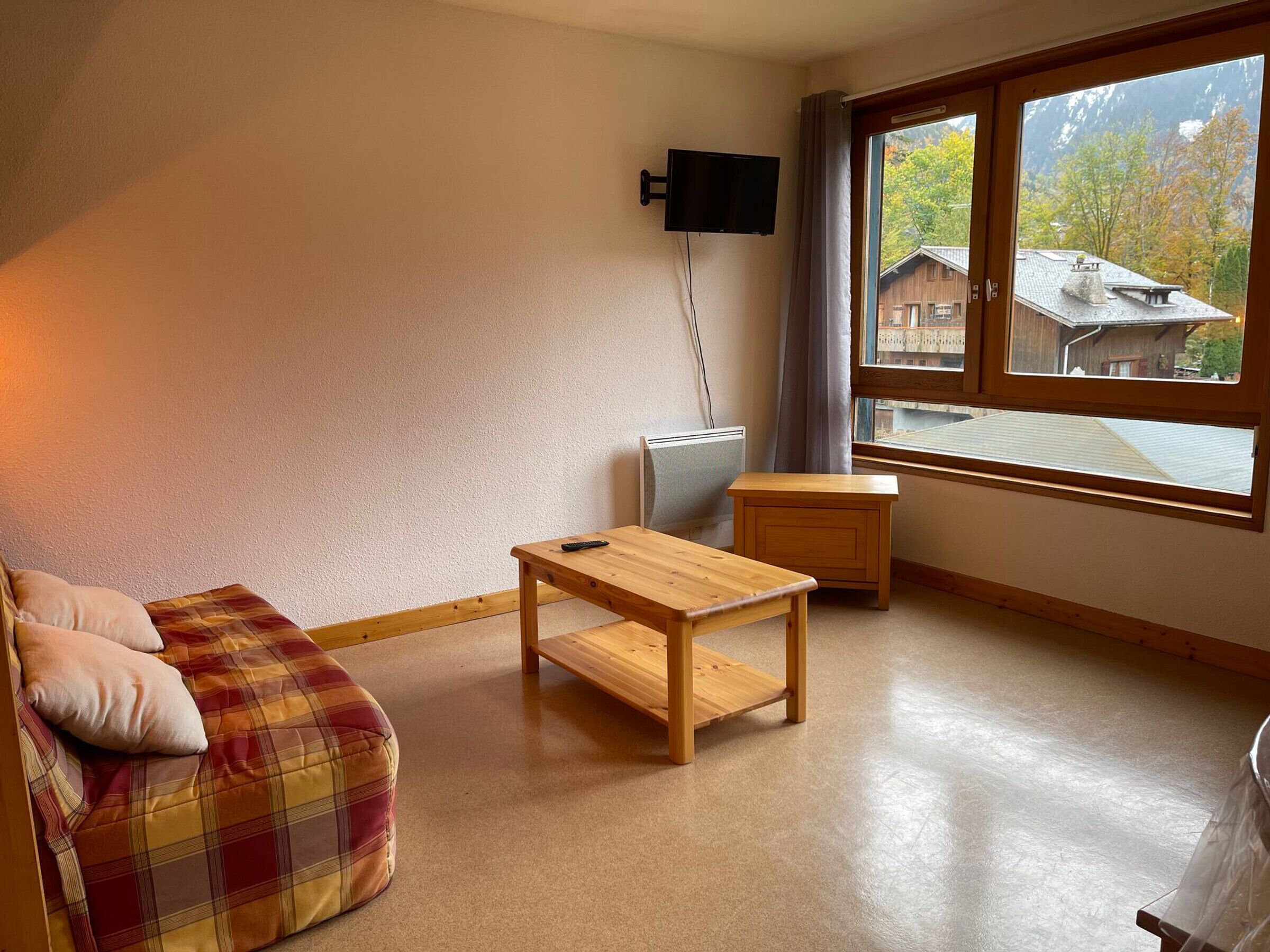 A cozy living room with a plaid sofa, a wooden coffee table, and a small side table. A flat-screen TV is mounted on the wall, and a window offers a view of a mountain and houses.