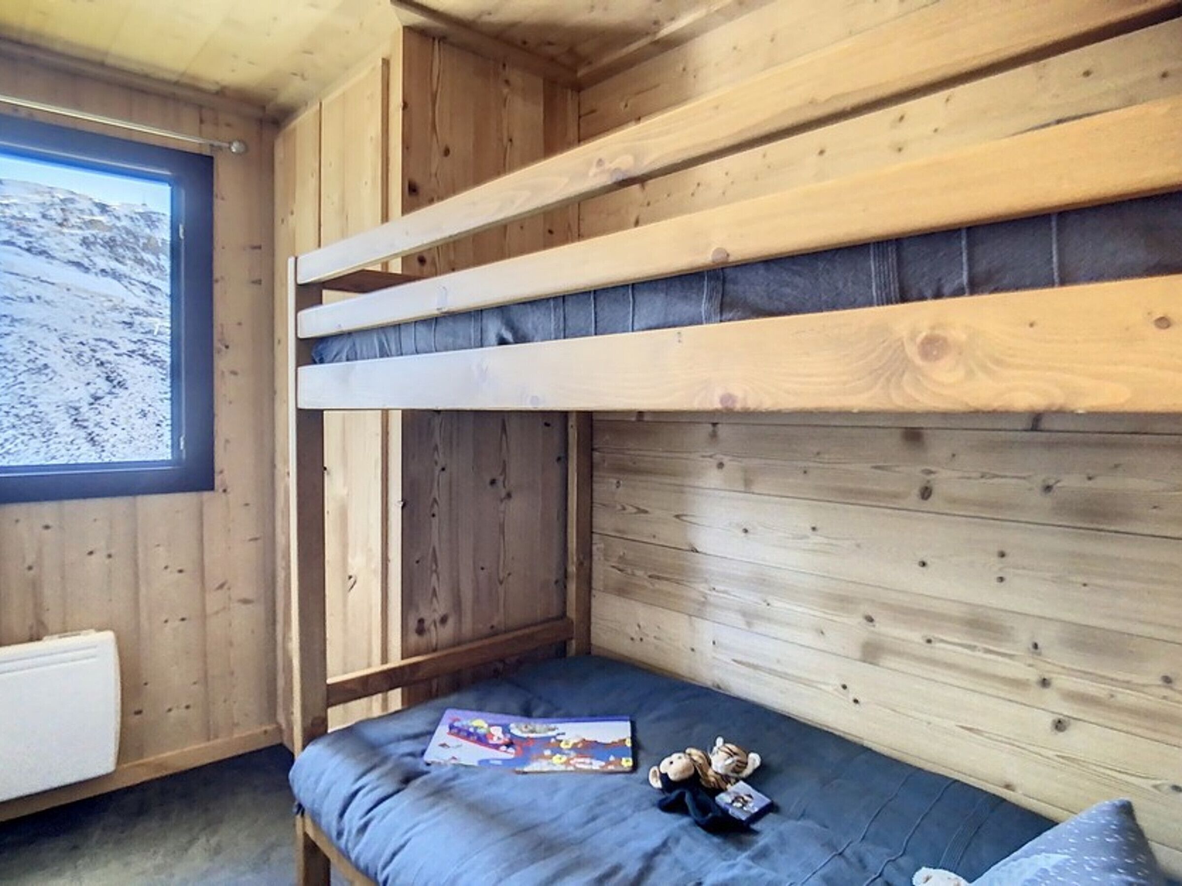 Un lit superposé avec une tête de lit en bois, une fenêtre donnant sur un paysage enneigé et une petite table de chevet avec un livre et un ours en peluche.