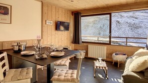 Et hyggeligt værelse med træloft, et spisebord dækket med tallerkener og glas, et fjernsyn på væggen og udsigt til et sneklædt landskab gennem vinduet.