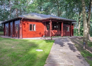 A wooden cabin with a porch, a stone pathway, and a grassy yard surrounded by trees. - Woodland Hawthorn Lodge (Ellesmere)