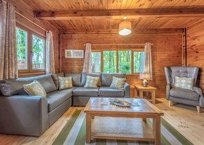 A wooden cabin interior with a sectional sofa, a chair, a table, and a lamp. - Woodland Hawthorn Lodge (Ellesmere)