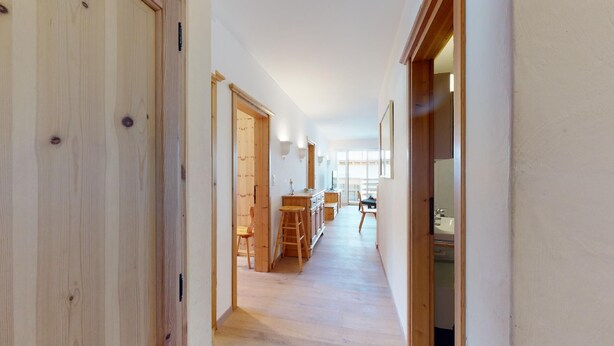 Un couloir étroit, doté de planchers en bois et de portes donnant sur un espace cuisine comprenant une table et des chaises.