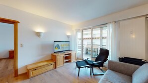 Un salon moderne avec un meuble de rangement en bois pour l’équipement audiovisuel, un fauteuil de bureau noir, un canapé gris et une grande fenêtre donnant sur un balcon.