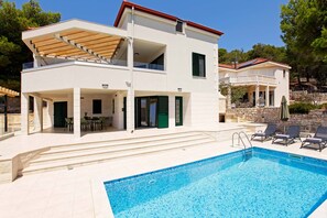 A two-story modern house with a swimming pool, outdoor seating, and a stone wall.