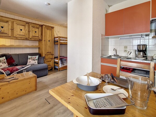 A cozy room with a bunk bed, a sofa, a wooden table with kitchenware, and a kitchenette with orange cabinets. - Cozy studio at ski slopes with balcony (Les Menuires)