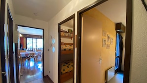 Un couloir mène à une chambre avec un lit superposé, un coin repas avec une table et des chaises, et une cuisine équipée d’un réfrigérateur.