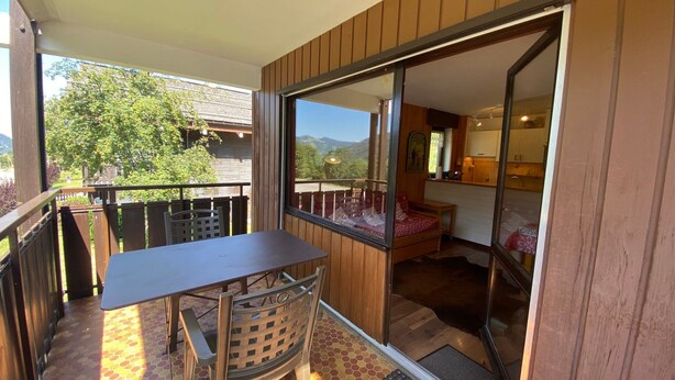 Un balcon avec une table et des chaises, une porte-fenêtre coulissante donnant sur une chambre, et une vue sur une montagne et des arbres.