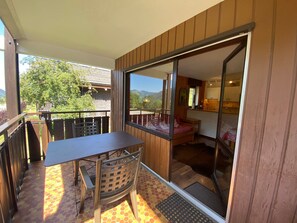 Un balcón con mesa y sillas, una puerta corrediza de vidrio que da a un dormitorio y vistas a una montaña y árboles.