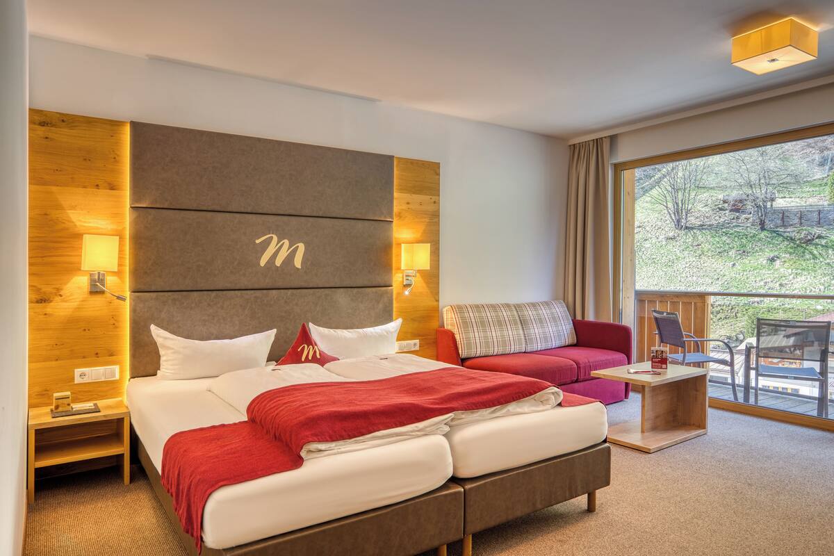 A modern hotel room with a large bed, a red sofa, a wooden headboard, and a balcony with outdoor furniture.