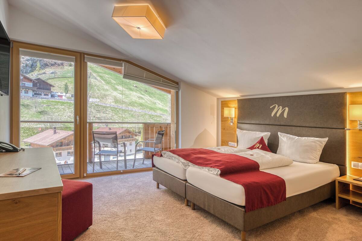 A modern hotel room with a large bed, a desk, and a balcony with a view of the countryside.