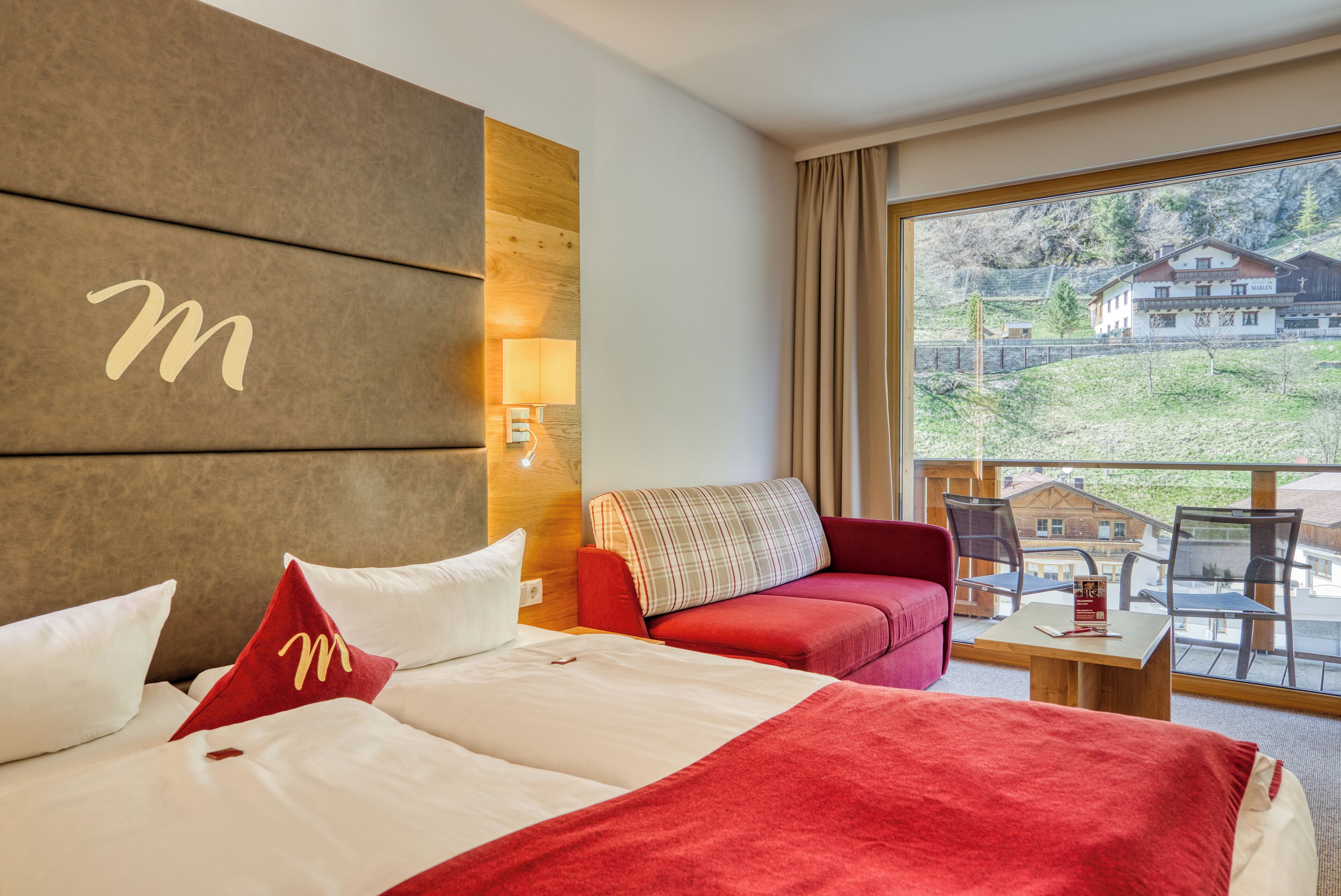 A hotel room with a large bed, a red sofa, a wooden headboard, a window with a view of houses, and a small table with a book.