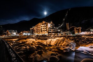 A multi-story hotel with a prominent sign, situated by a river with rocks and snowbanks. - Hotel Erlebnis Mallaun (See)