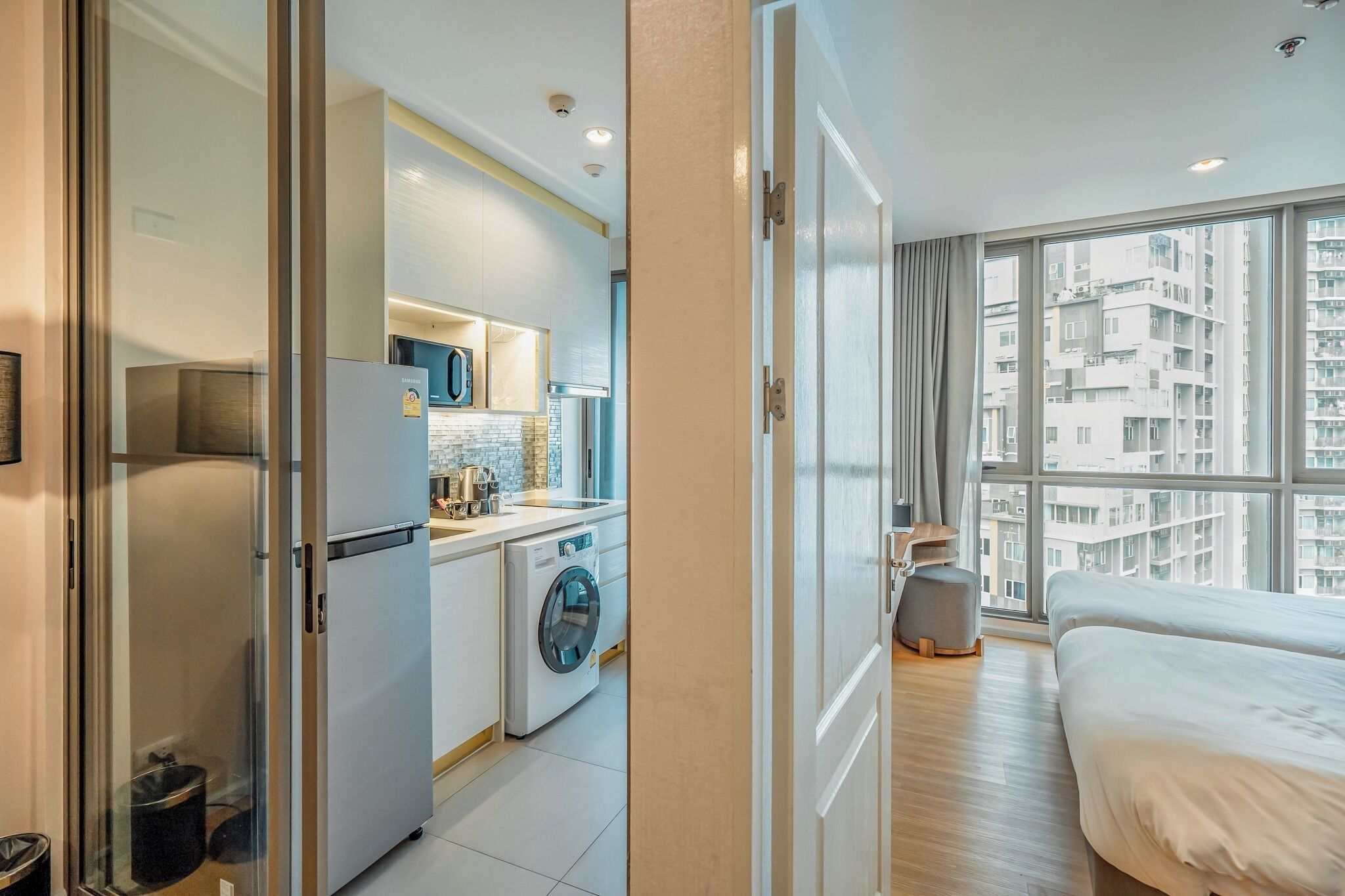 A modern apartment with a kitchen, a washing machine, a bed, and a view of high-rise buildings.