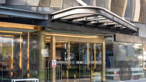 Una entrada moderna de edificio con puerta de vidrio, estructura metálica y un banco en el exterior.