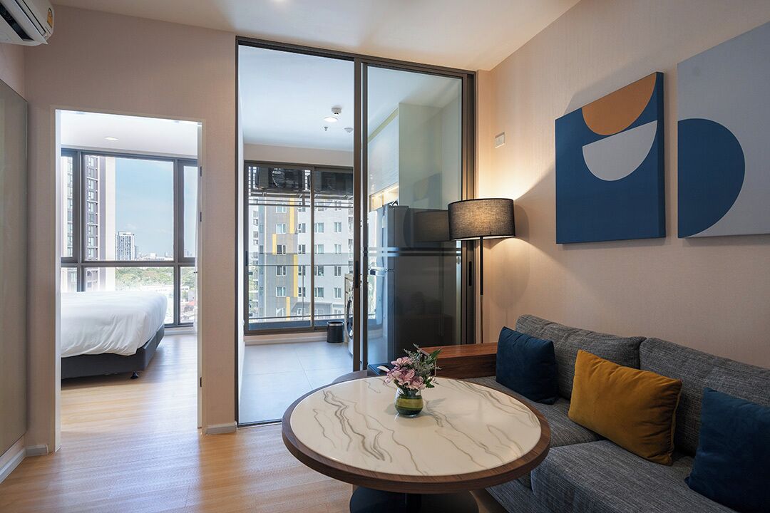 Quarto de hotel moderno com cama, mesa redonda com um vaso, sofá com almofadas e uma grande janela com vista para a cidade.