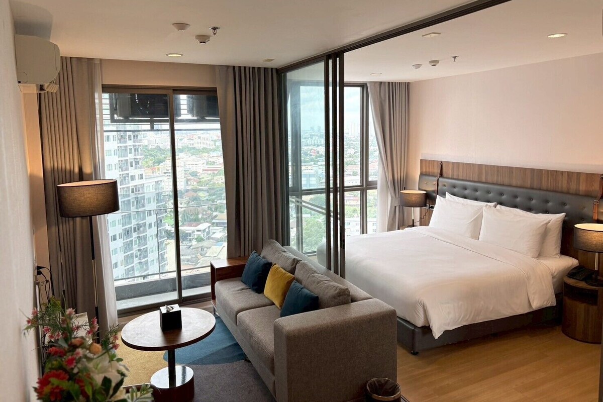 Ein modernes Hotelzimmer mit einem großen Bett, einer Couch, einem kleinen Tisch und Blick auf die Stadt durch große Fenster.