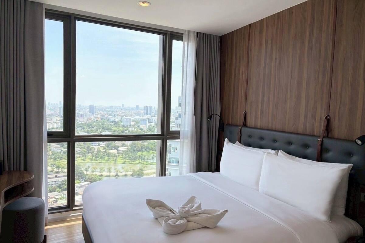 Een hotelkamer met een groot raam, een bed met witte lakens, een houten hoofdeinde en uitzicht op een stadsgezicht.