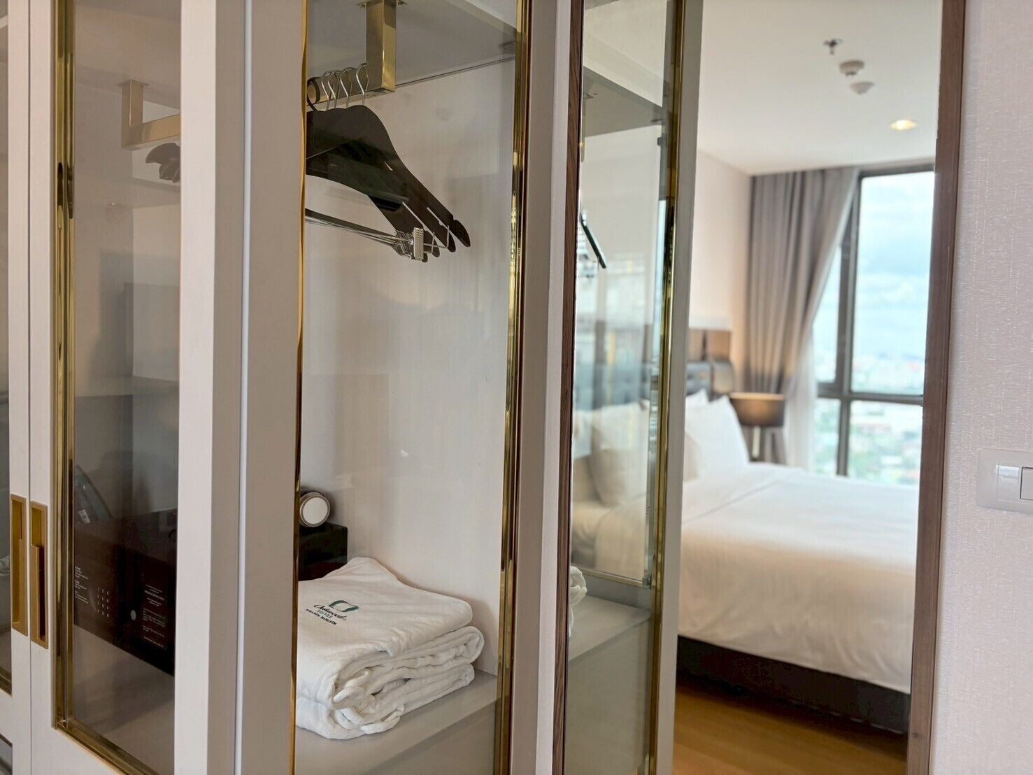 Une chambre d’hôtel dotée d’un dressing fermé par des parois de verre, d’un lit parfaitement fait et offrant une vue sur la ville par la fenêtre.