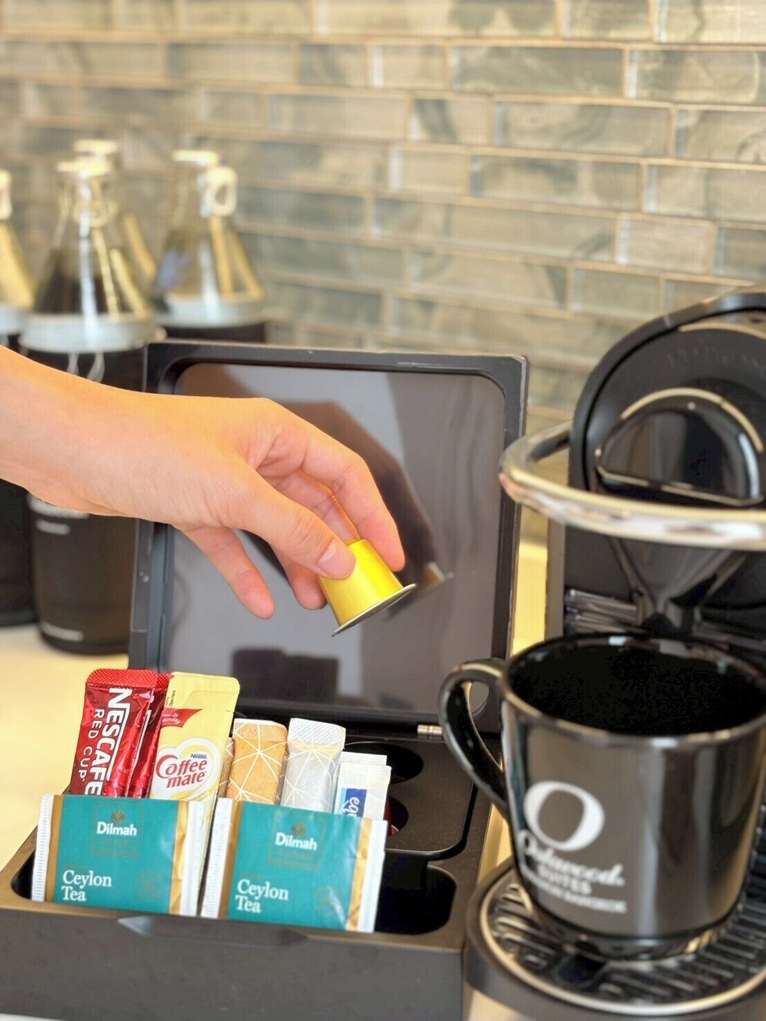 A coffee machine with a hand holding a coffee pod, a coffee pot, and a tray with various coffee pods and a tea bag.