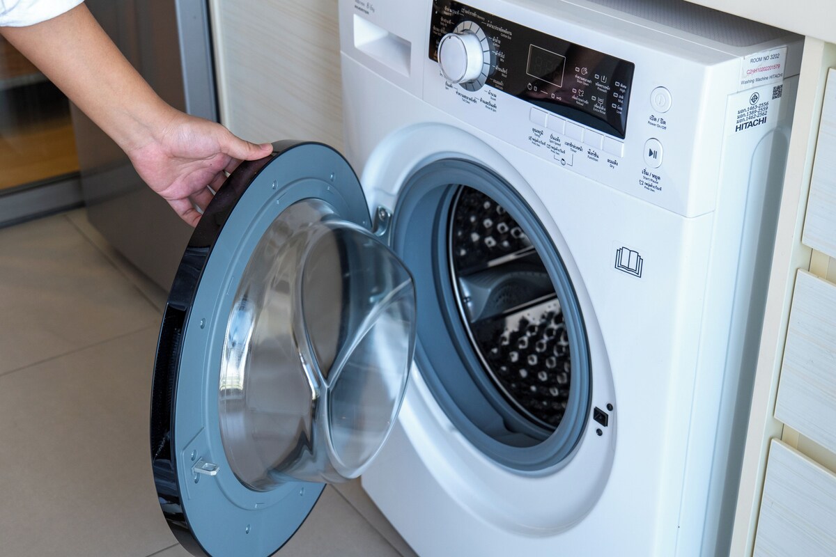 Eine Waschmaschine mit Frontlader, ein Bedienfeld und eine Hand, die die Tür öffnet, mit dem Hinweis 'ZIMMER NR. 3022'.