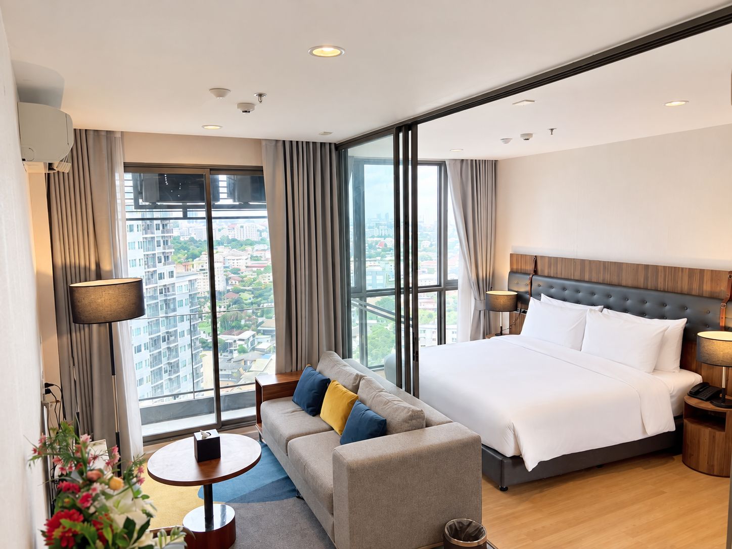 A modern hotel room with a large bed, a sofa, a small table, and a view of the city through large windows.