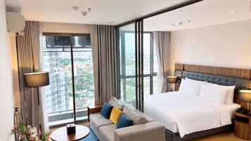 A modern hotel room with a large bed, a sofa, a small table, and a view of the city through large windows.