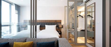 Habitación de hotel moderna con una cama grande, un sofá gris con cojines, un vestidor y un ventanal con vistas.