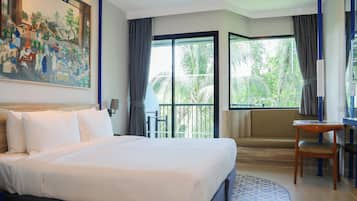 A hotel room with a large bed, a desk, a chair, a painting on the wall, and a balcony with a view of palm trees.